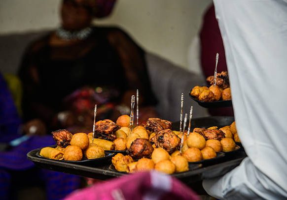 Server presenting assorted Nigerian small chops platter, featuring puff-puff, grilled chicken, samosas, and spring rolls at a social event in Lagos